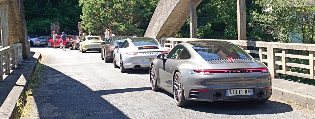 Une file de voitures de sport Porsche, principalement grises et blanches, est garée sur un pont ensoleillé, avec des gens debout à proximité et des arbres en arrière-plan.