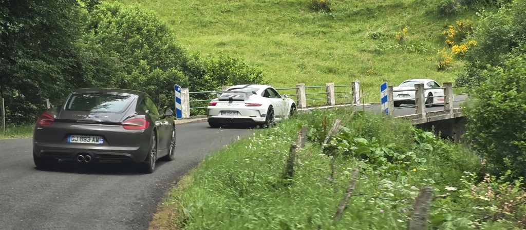 Trois voitures de sport, dont une décapotable noire et deux coupés blancs, roulent sur une route de campagne étroite et sinueuse, bordée de verdure et d'un petit pont aux balises bleues et blanches.