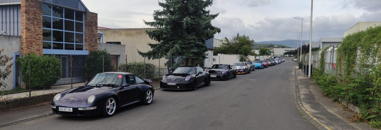 Une rangée de voitures de sport, principalement des modèles Porsche, est garée le long d'une rue tranquille bordée de bâtiments industriels et de verdure sous un ciel nuageux.