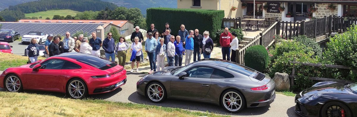 IMG_20250727_124441b Un groupe de personnes se tient à l'extérieur d'un bâtiment, près de voitures de sport garées, dont une Porsche rouge et une Porsche grise, par une journée ensoleillée, avec des collines verdoyantes et des panneaux solaires en arrière-plan.