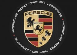 L'image représente le logo Porsche avec un cheval noir, des bandes rouges et noires et des bois. Autour, "Porsche Road Trip en Lorraine" est inscrit dans un cercle sur fond noir, célébrant ainsi les clubs indépendants et leur passion pour la conduite.